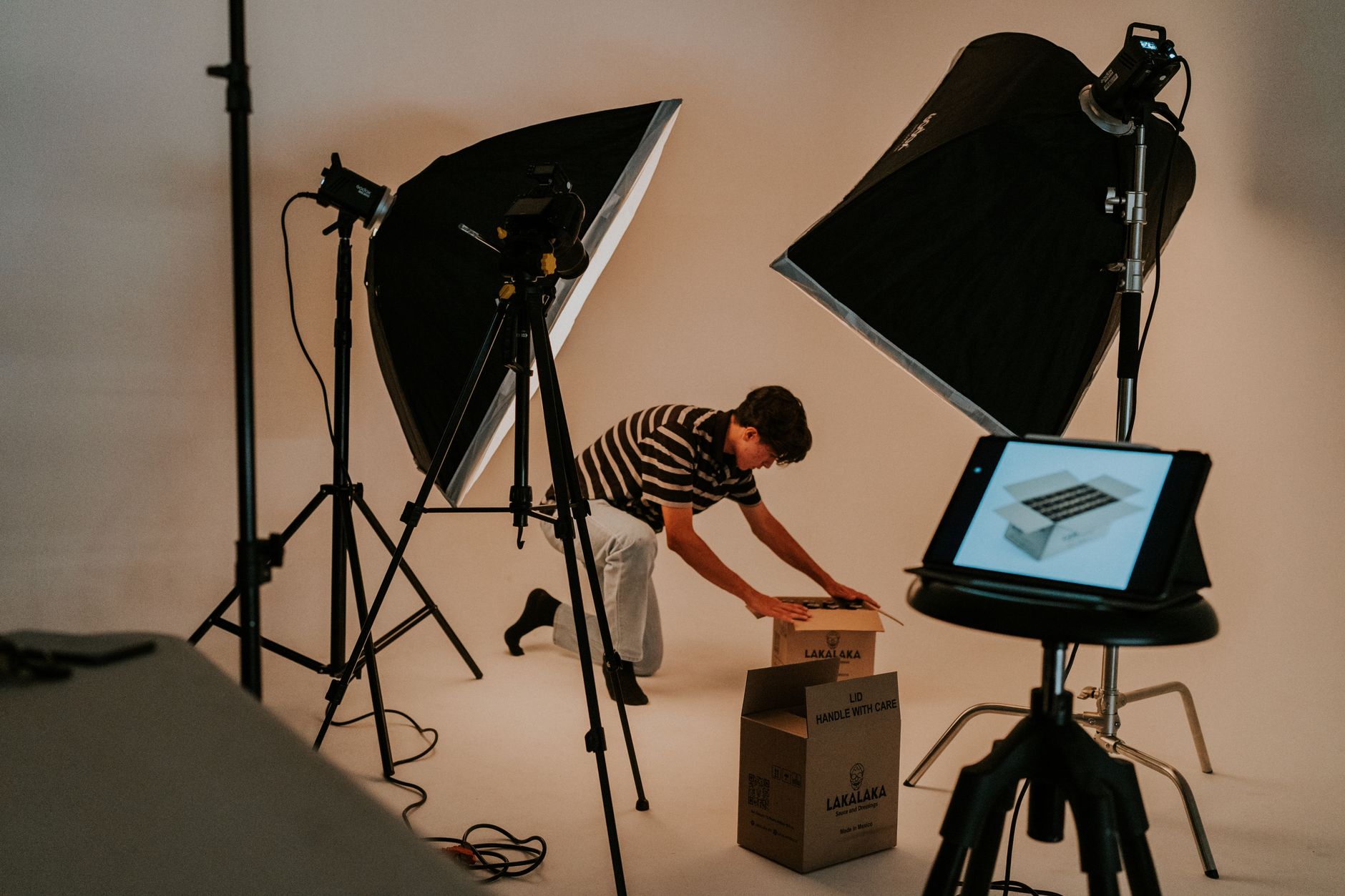 Un homme prépare un colis au centre d’un studio, entouré de lumières, de trépieds et d’une tablette
